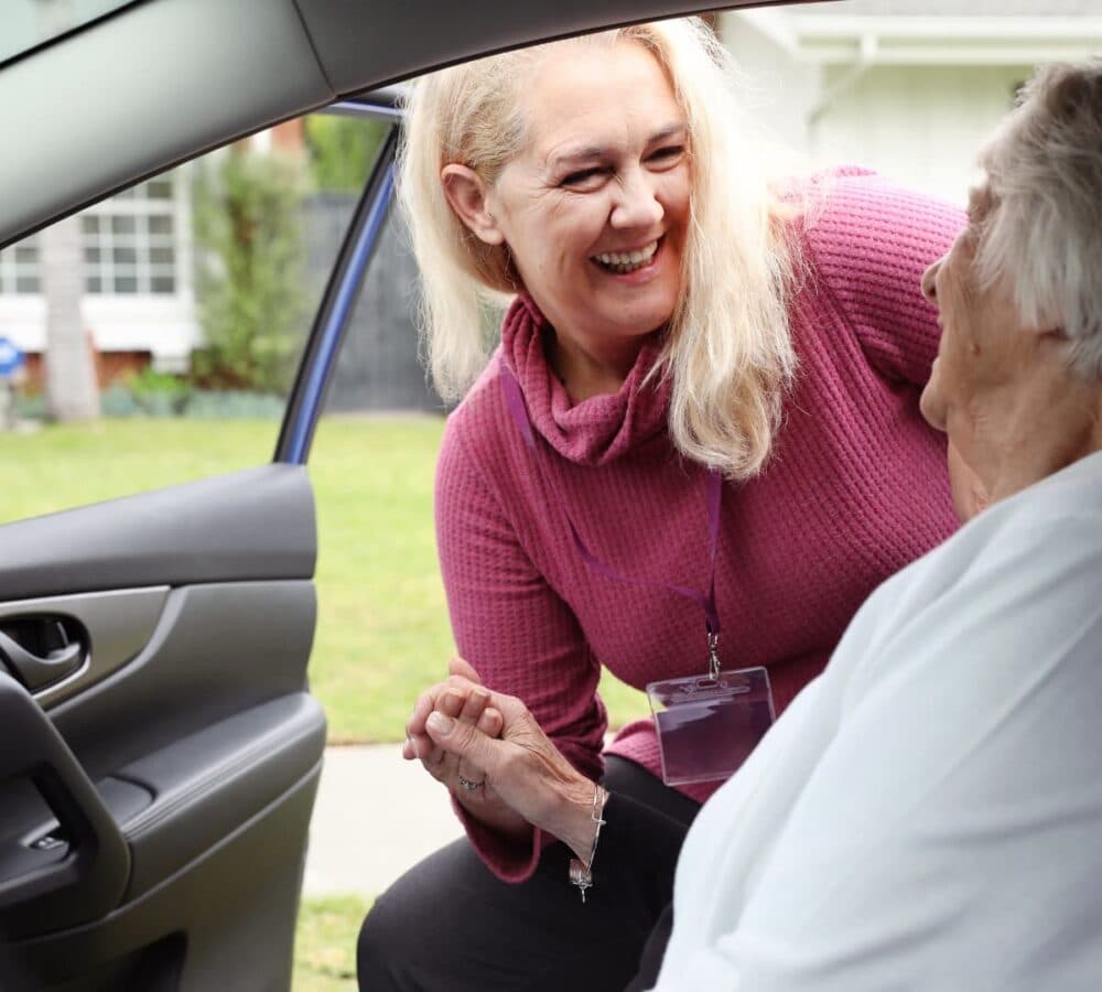 A young female carer with long hair and wearing pink sweater helping an ilder female adult to go out of the car