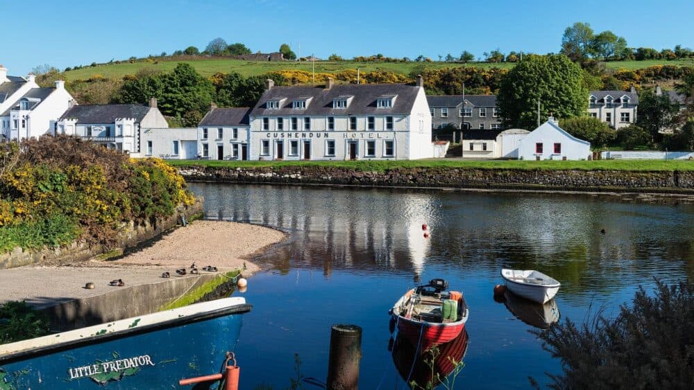 Boats on a calm river with the Cushendun Hotel and white houses in a green, hilly landscape. - Home Instead