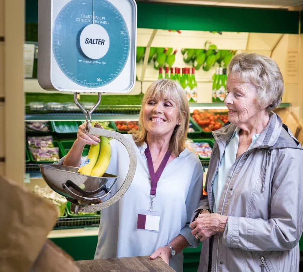 Younger woman with short hair weighing a banana with an older woman with short hair both smiling inside the grocery store