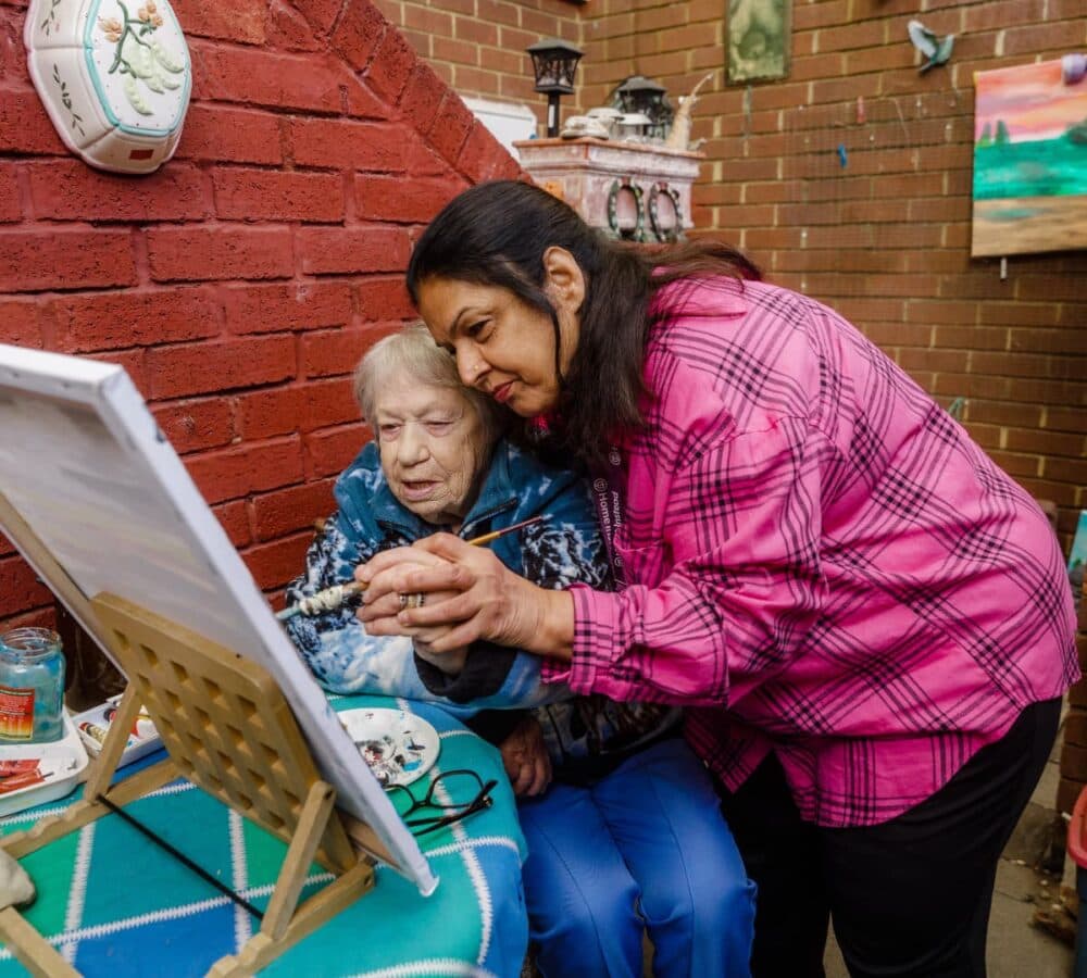An older female adult painting outside with her younger female carer with black hair and wearing pink