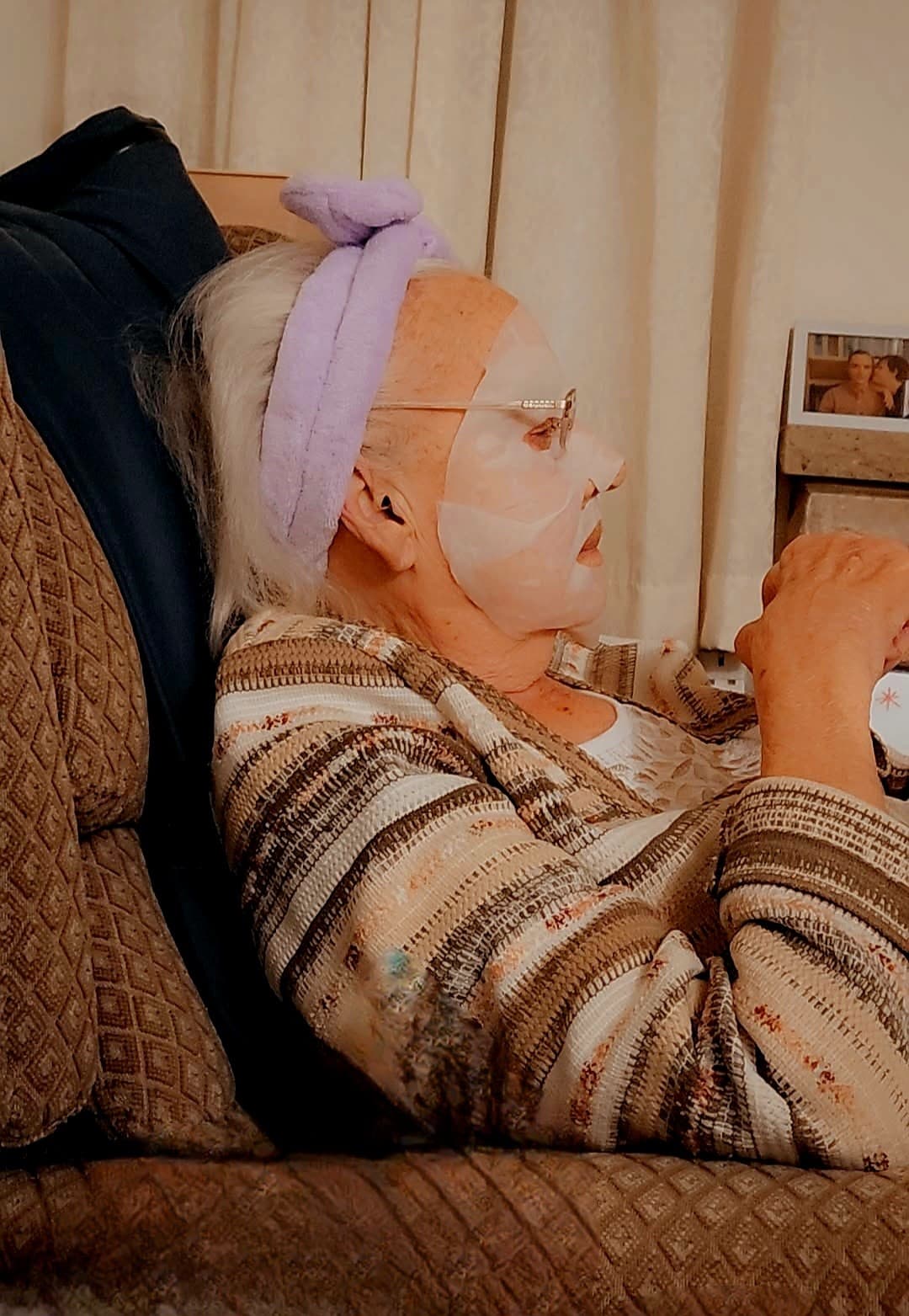 Elderly woman lying on a couch wearing a face mask and headband, holding a cup. - Home Instead