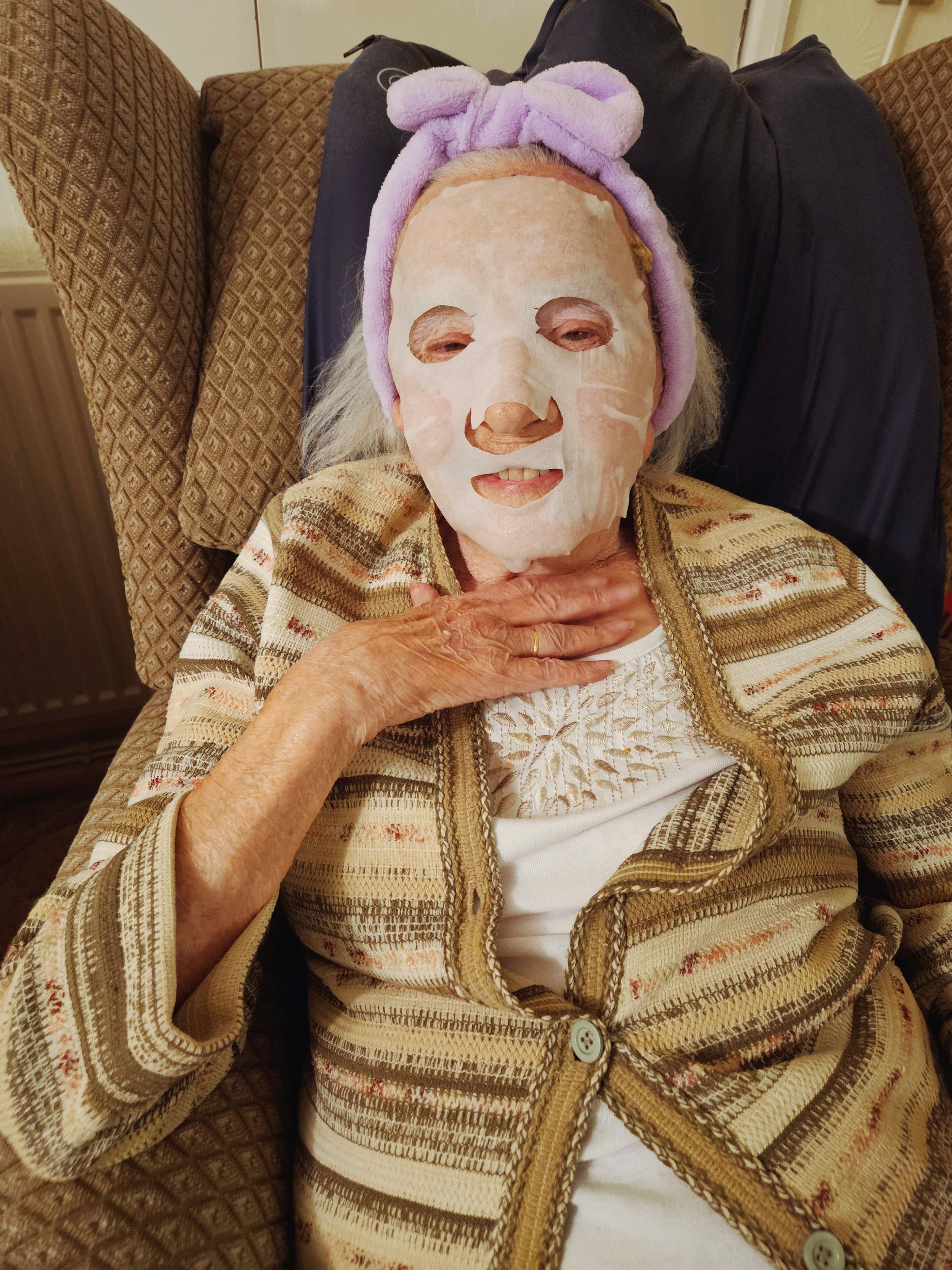 Elderly woman wearing a face mask and a purple headband, sitting in an armchair with hand on her chest. - Home Instead