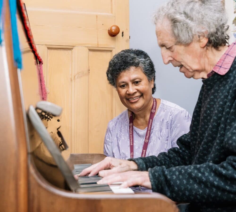 An older male adult with grey hair playing the piano with his younger female carer watching him