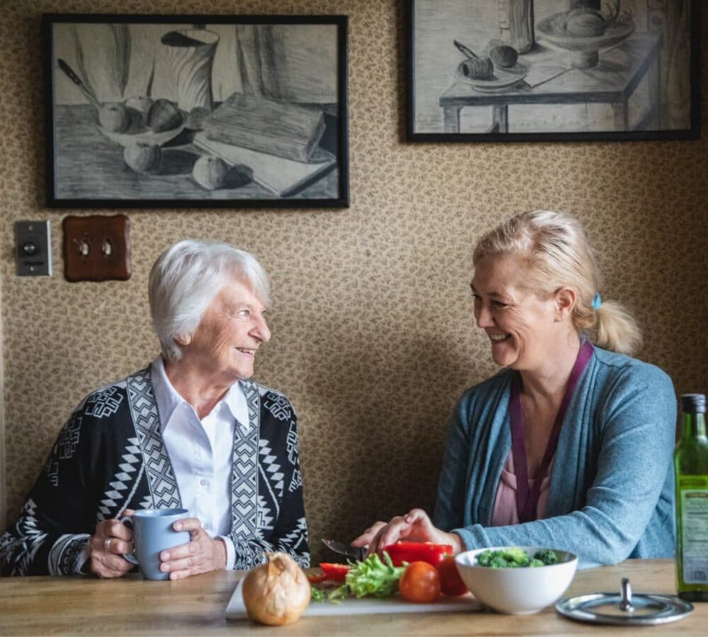 Younger female carer cutting vegetables with an older woman with grey hair holding a mug both happy and smiling