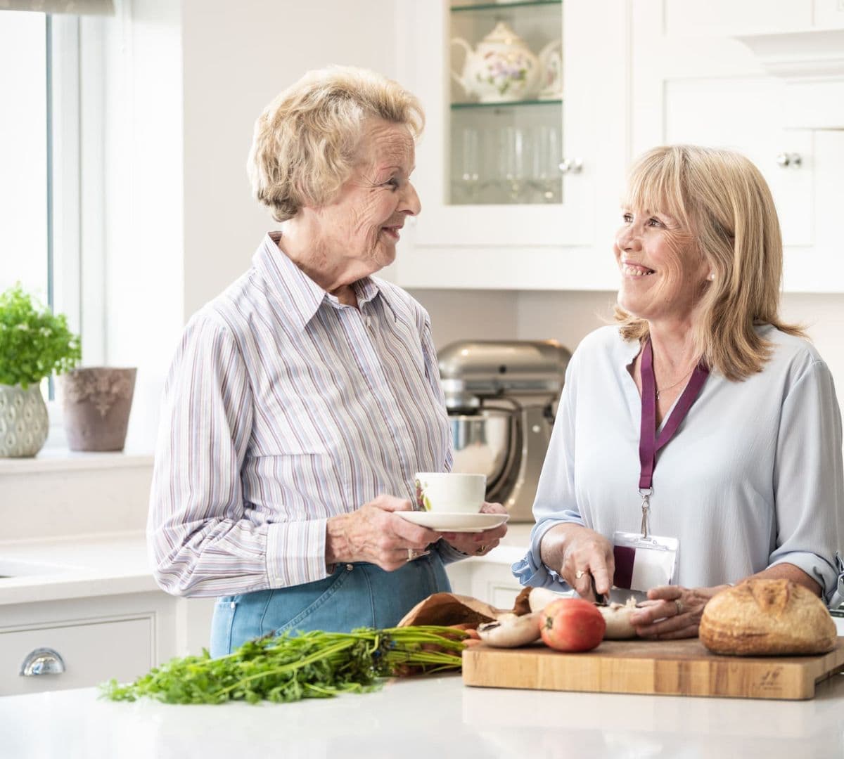 A younger carer cutting vegetables inside the kitchen with an older female adult with short hair holding a cup both happy and smiling