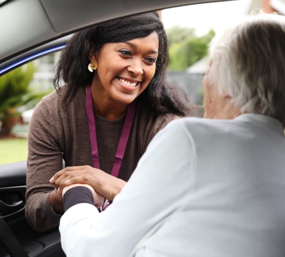 A woman with black hair smiling at the door of the car helping an older adult woman getting out of the car