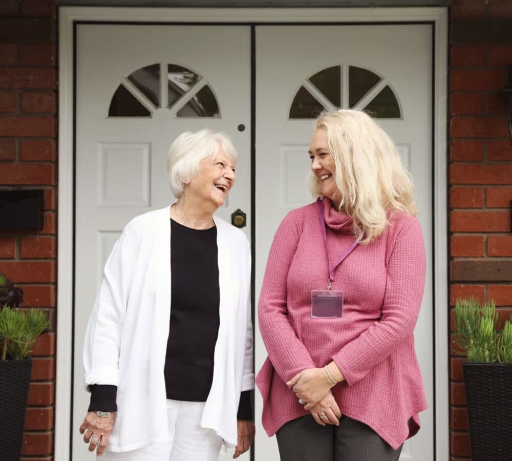 Two women both happy and smiling, younger woman with blonde hair and wearing pink and an older woman with short hair and wearing white