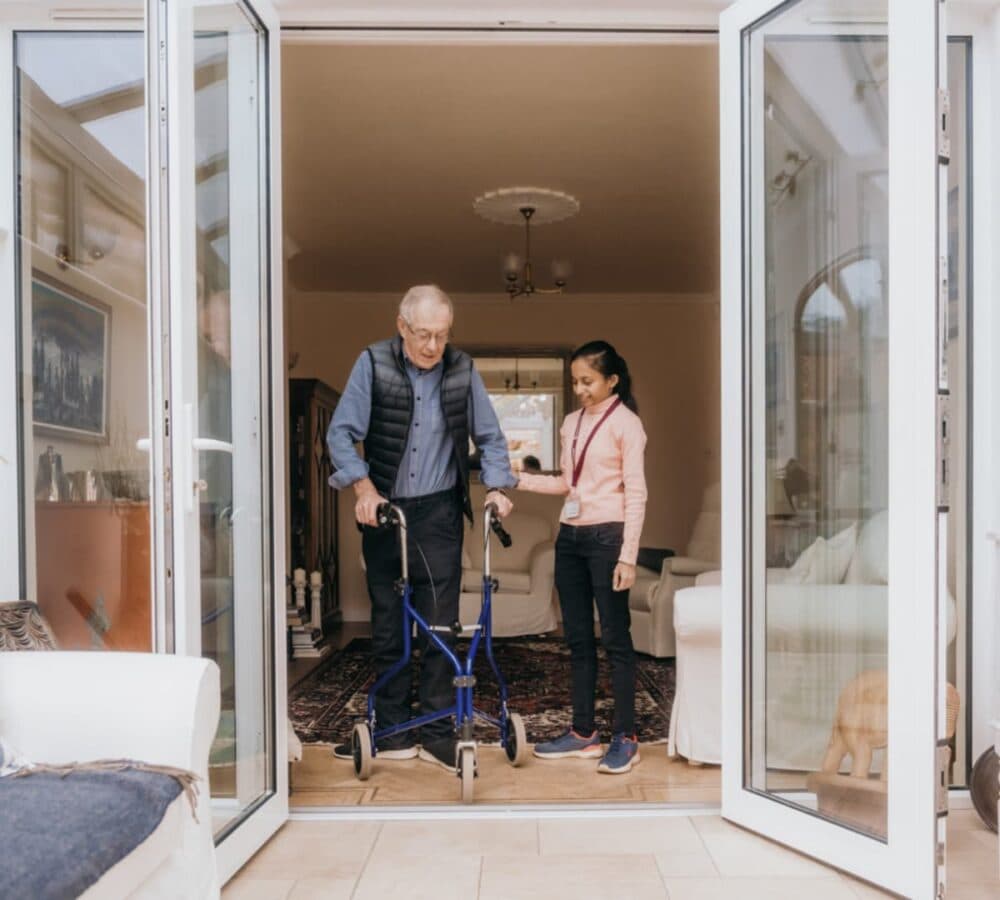 A male older adult walking out the door while using a walker and being helped by his younger female carer