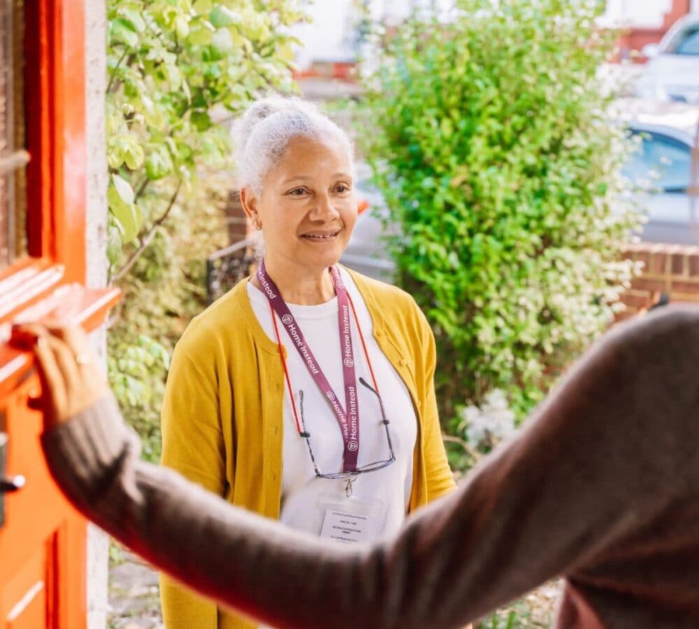 Woman standing at the door wearing yellow sweatshirt and with grey hair and smiling