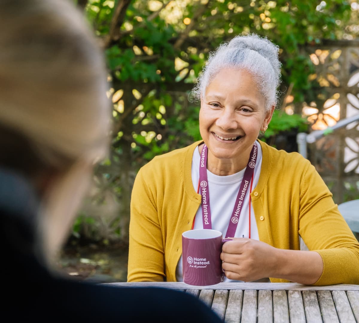 An older woman with grey hair smiling and happy while holding a Home Instead mug outdoors with green leaves in the background