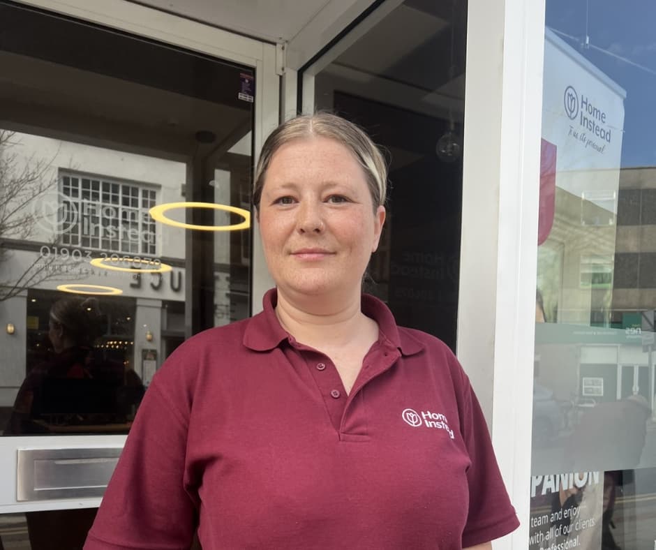 A woman in a maroon Home Instead polo shirt stands outside a building, looking at the camera. - Home Instead