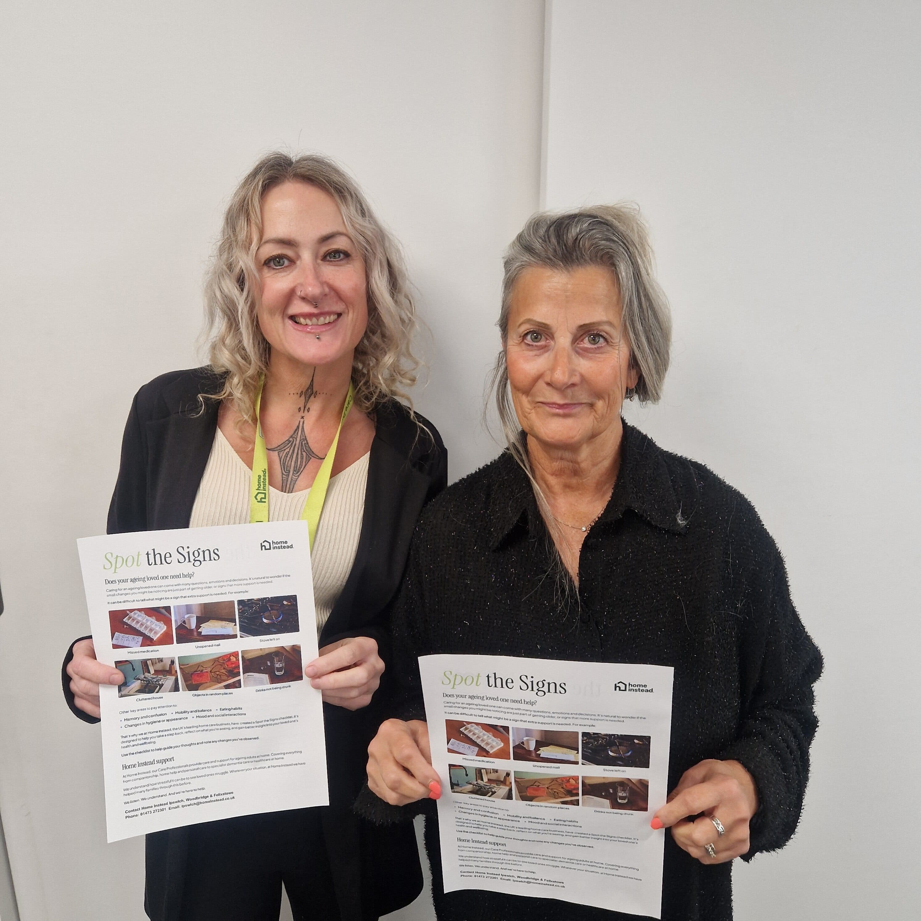 Two women smiling and holding "Spot the Signs" information sheets against a plain white wall. - Home Instead