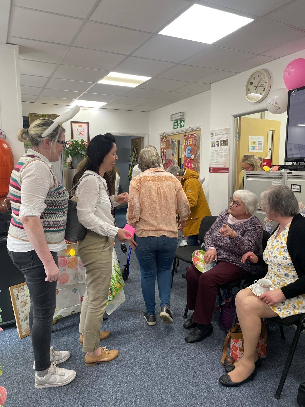 A group of women chat and interact at a cheerful indoor social gathering with decorations. - Home Instead