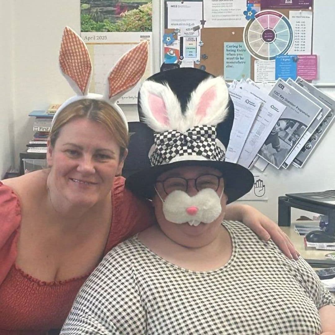 Two women smiling in an office, one wearing bunny ears and the other in a hat with bunny ears and a fake nose. - Home Instead