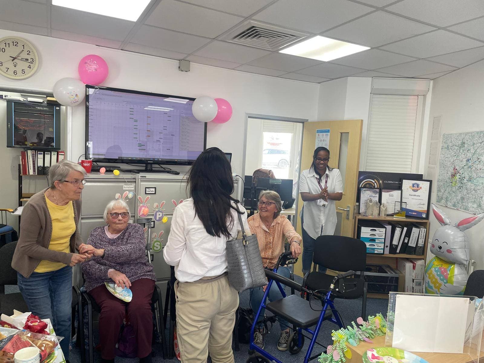 Three elderly women, a staff member, and a visitor chat in a decorated office with balloons and festive decor. - Home Instead