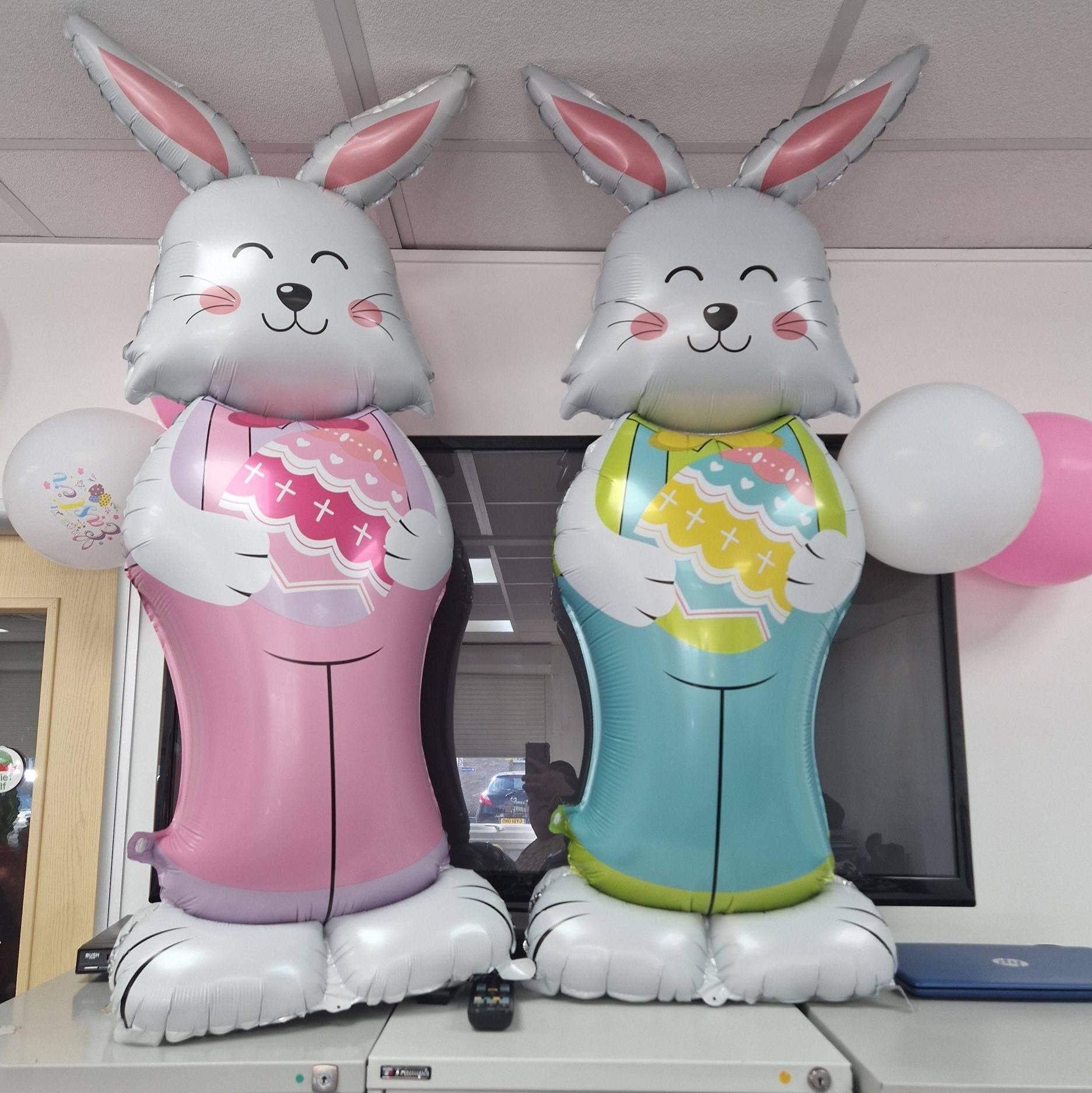 Two large bunny balloons, one pink and one blue, each holding a decorated Easter egg, with balloons in the background. - Home Instead