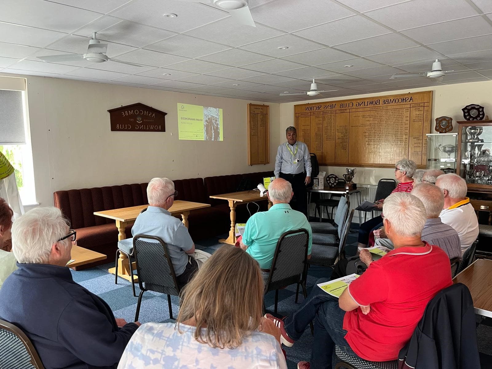 A group of seniors listen to a man giving a presentation in a club room with wooden honor boards. - Home Instead