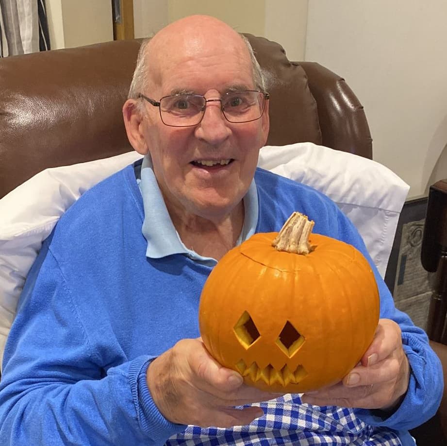Elderly man smiling and holding a carved pumpkin with geometric eyes and a jagged mouth, seated in a chair. - Home Instead