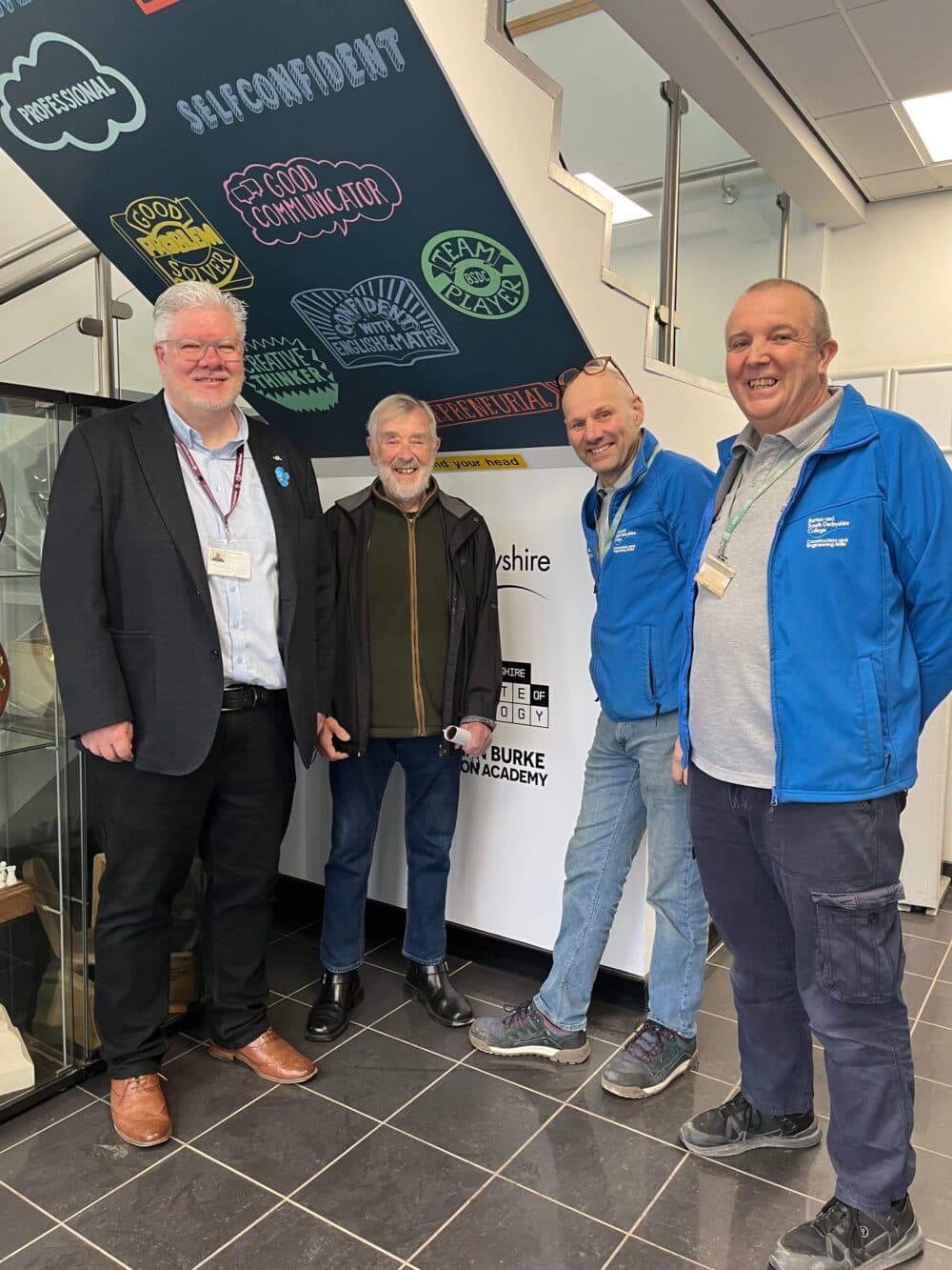 Four smiling men stand indoors near a wall with motivational words and icons under a staircase. - Home Instead