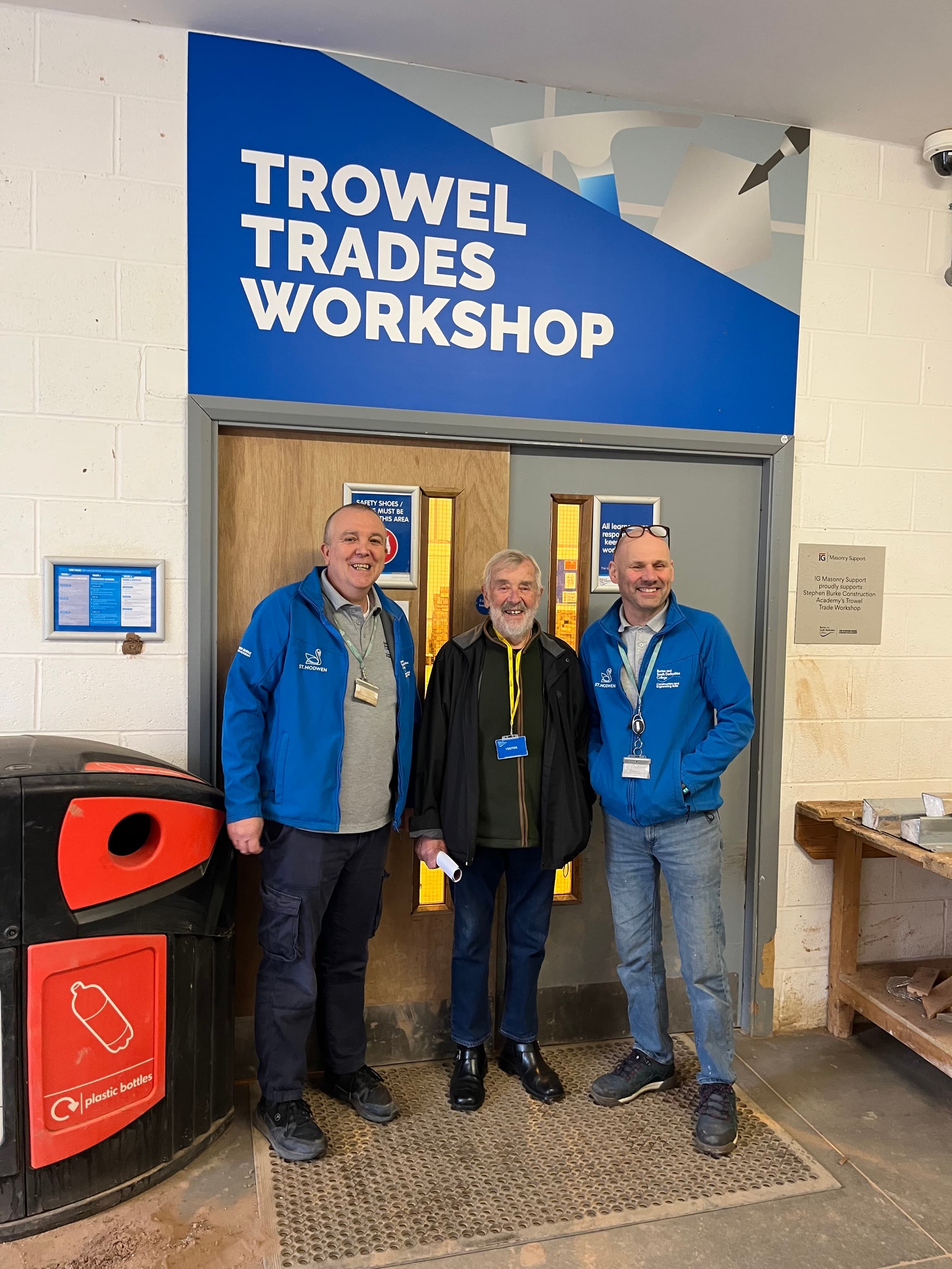 Three men stand smiling in front of a door labeled "Trowel Trades Workshop" inside a building. - Home Instead