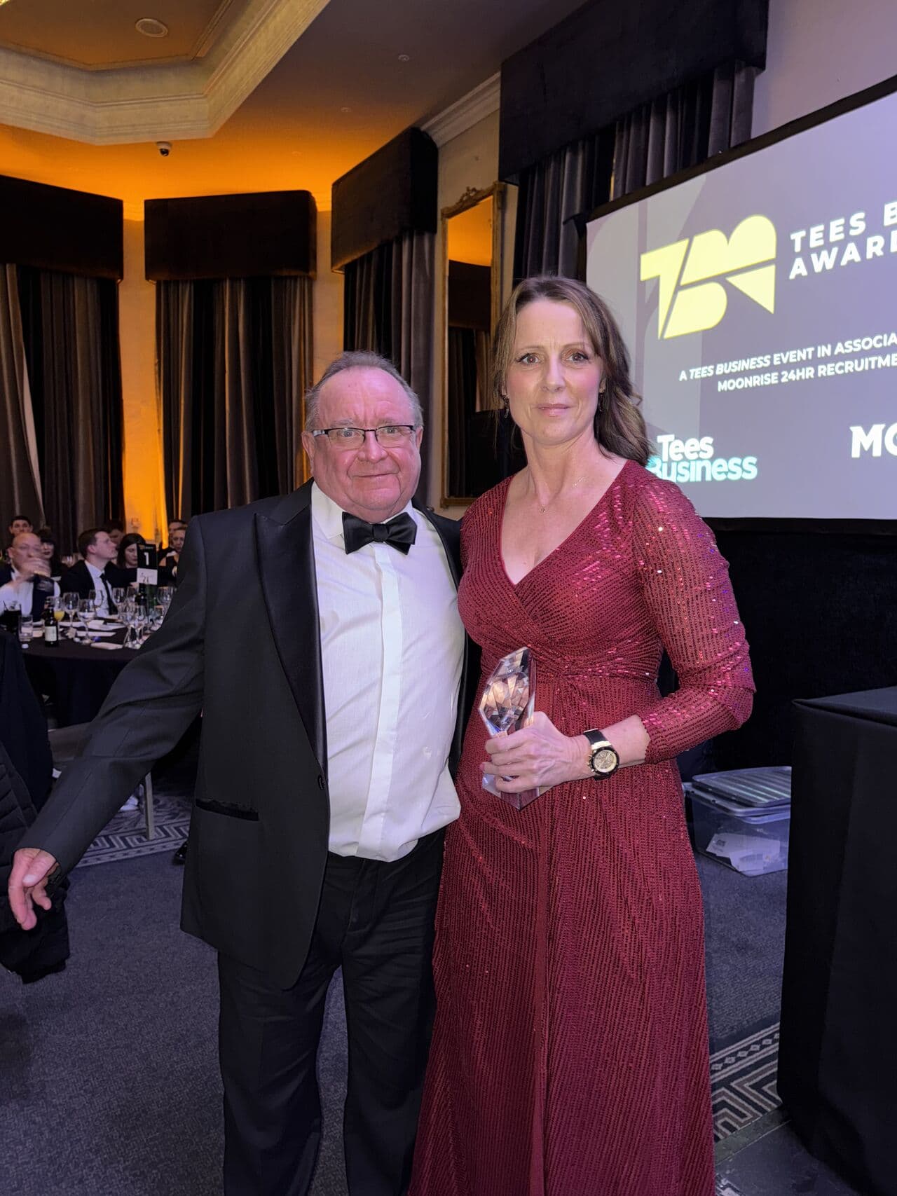 A man in a tuxedo and a woman in a red dress holding an award stand together at an indoor awards event. - Home Instead