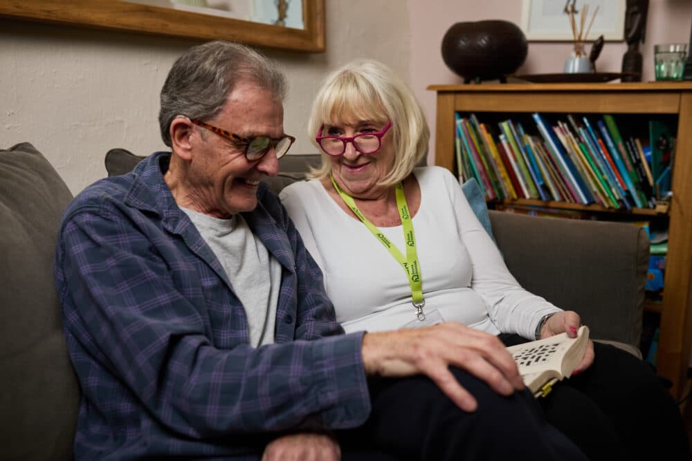 An elderly man and woman sit on a couch together, smiling and reading a book. - Home Instead