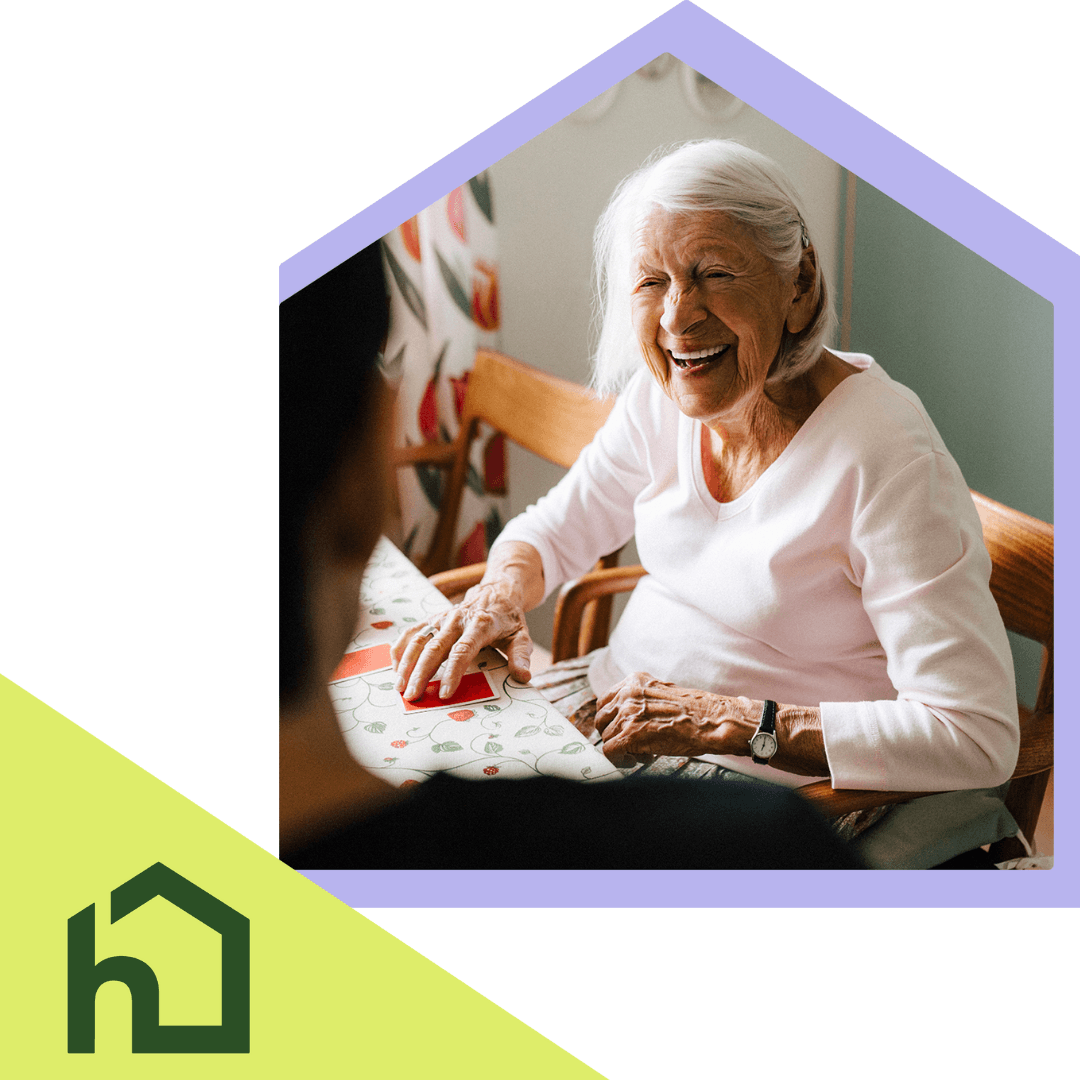 Smiling elderly woman sits at a table, talking with someone in a bright, cheerful room. - Home Instead