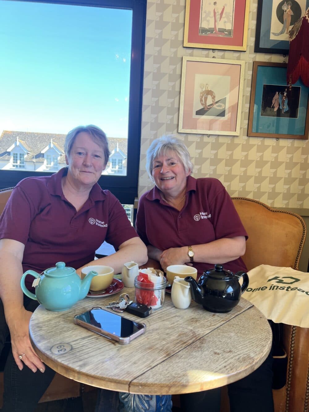 Two women in maroon shirts sit at a café table with tea, cups, and pastries, smiling at the camera. - Home Instead