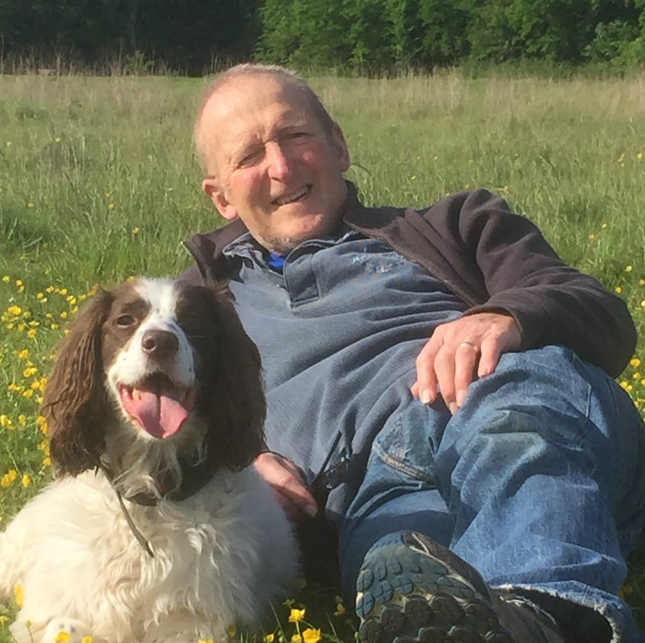 A man lies in a field of grass and yellow flowers, smiling with a happy dog by his side. - Home Instead