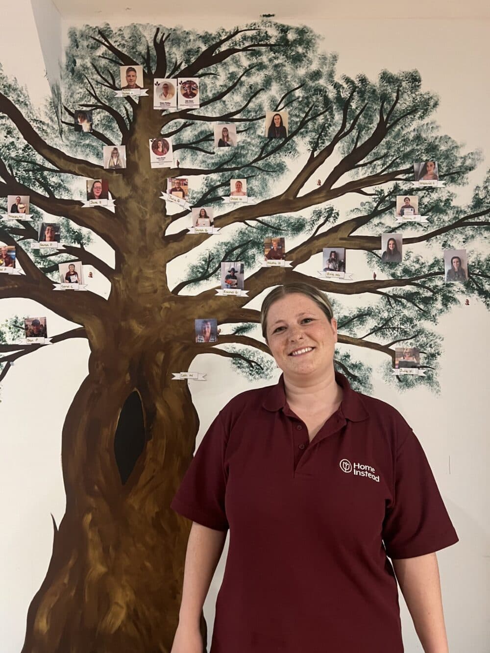 Woman in maroon uniform stands smiling in front of a painted family tree with photos on a wall. - Home Instead