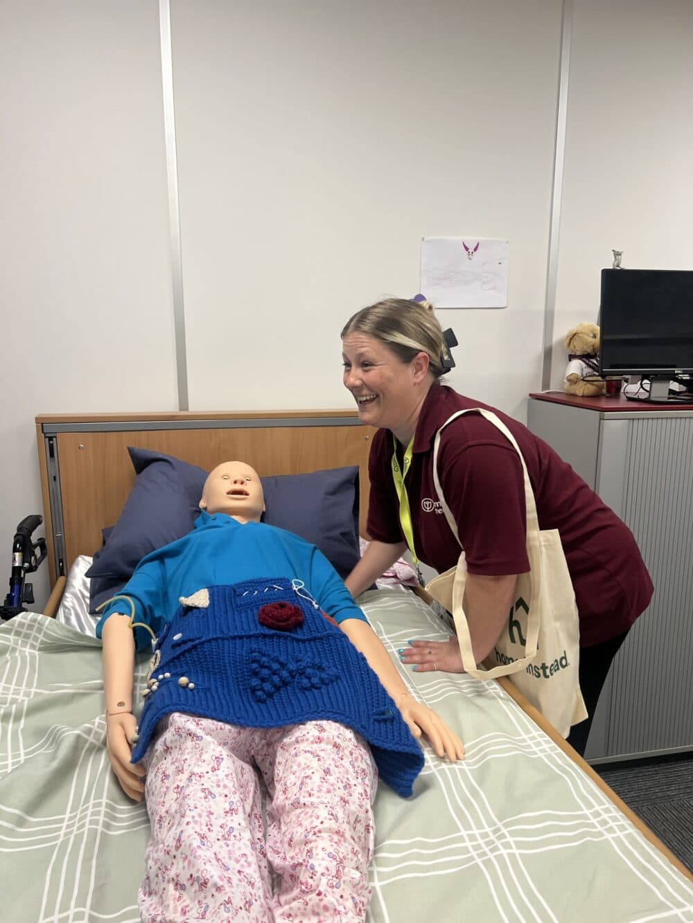 A woman smiles beside a medical mannequin lying in a hospital bed with a knitted blanket. - Home Instead