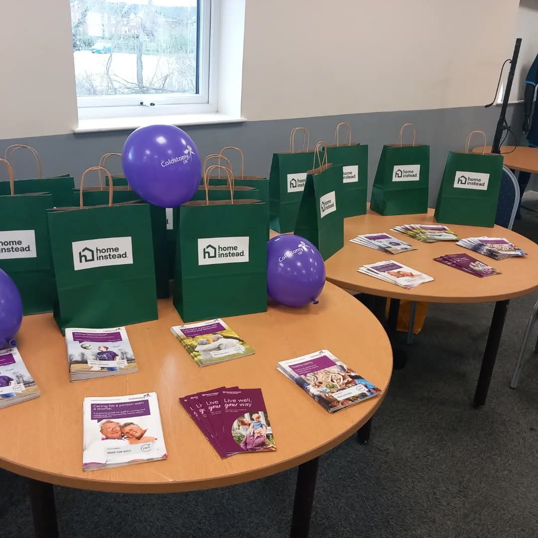 Two tables with green gift bags, purple balloons, and brochures arranged for a Home Instead event. - Home Instead