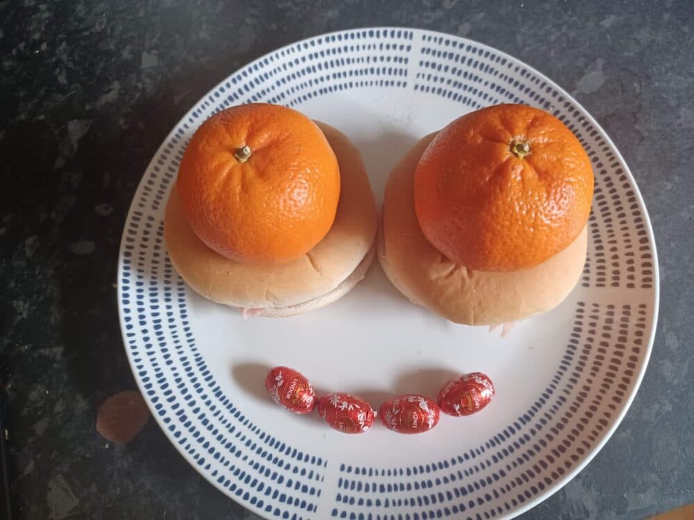 A plate with buns topped with oranges and five chocolate eggs arranged like a smiling face. - Home Instead