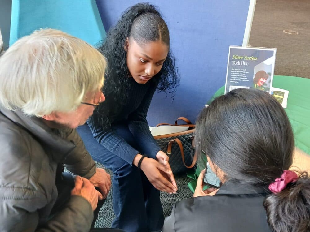 Young woman helps two older adults with a smartphone at a "Silver Surfers Tech Hub" event. - Home Instead