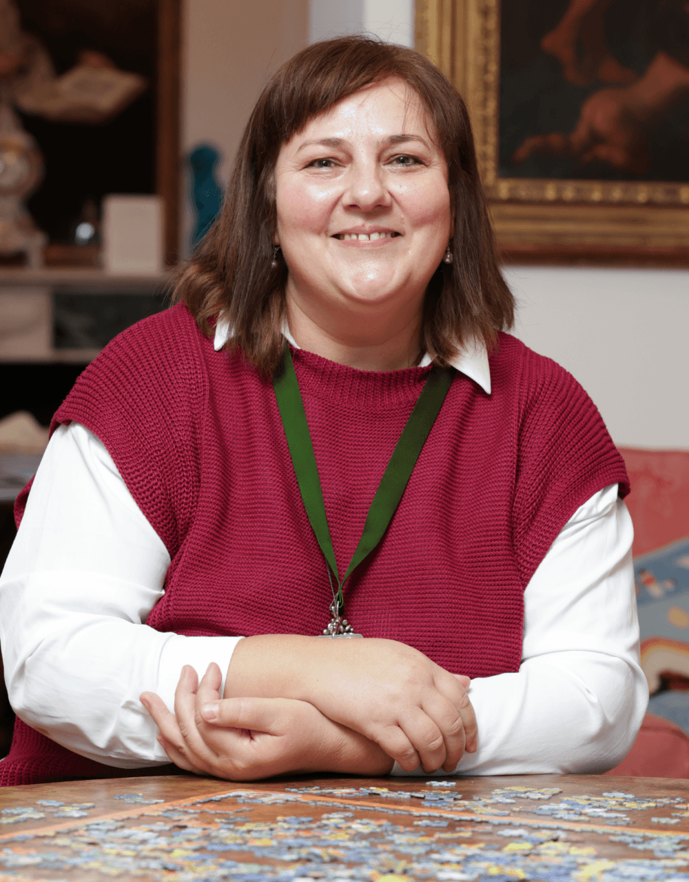 Smiling woman with brown hair wearing a red sweater sits at a table with a jigsaw puzzle. - Home Instead