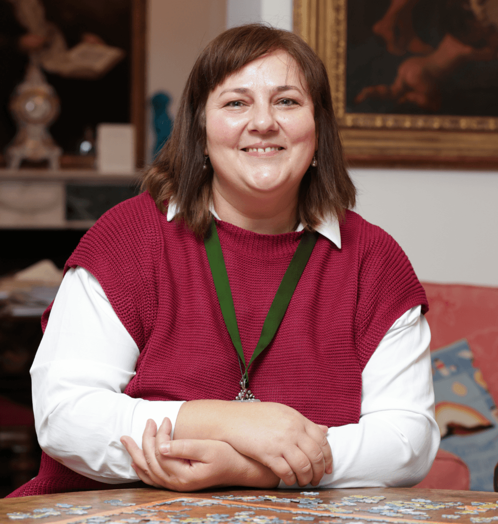 A woman in a red sweater sits at a table, smiling, with a puzzle in front of her in a cozy room. - Home Instead