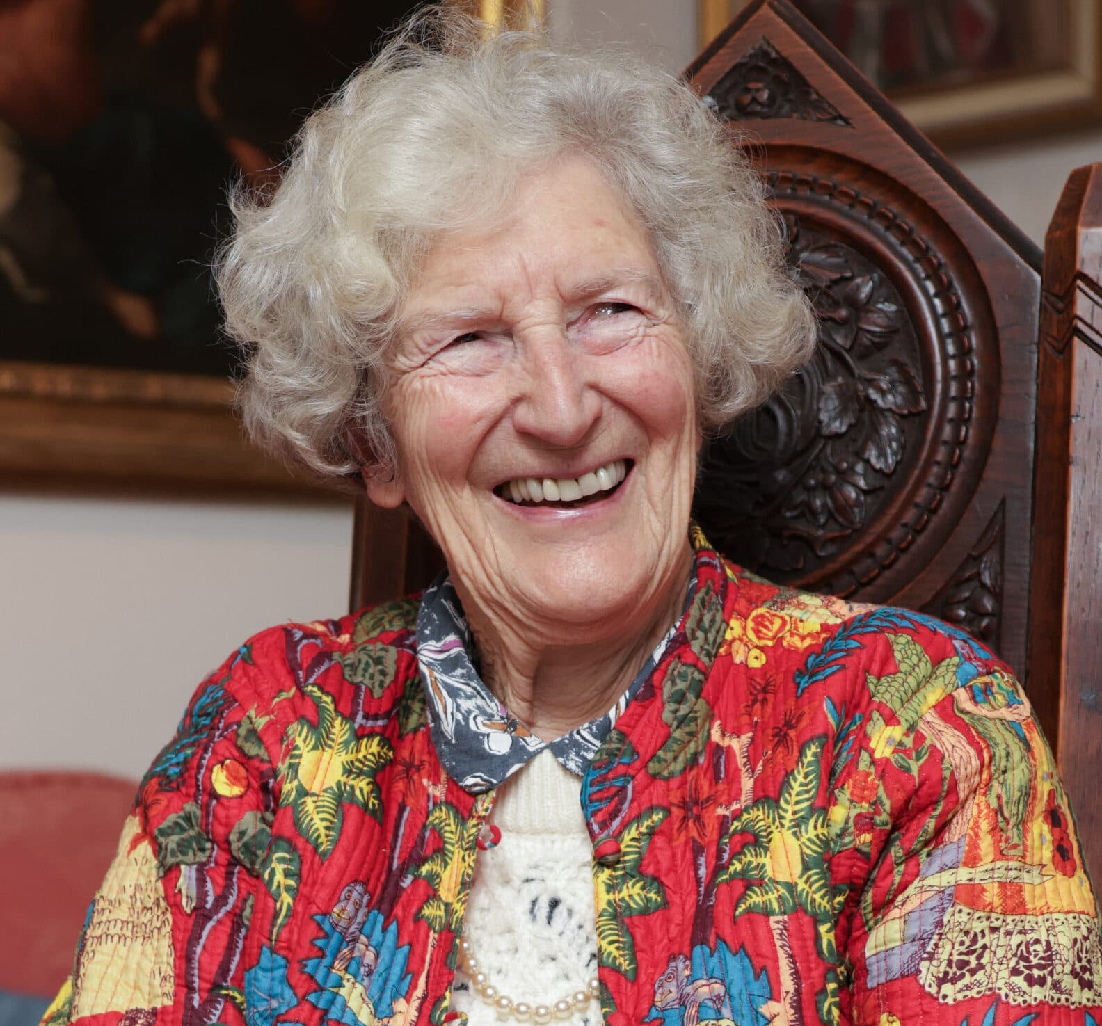Smiling elderly woman with curly gray hair in a colorful jacket, sitting on a carved wooden chair. - Home Instead