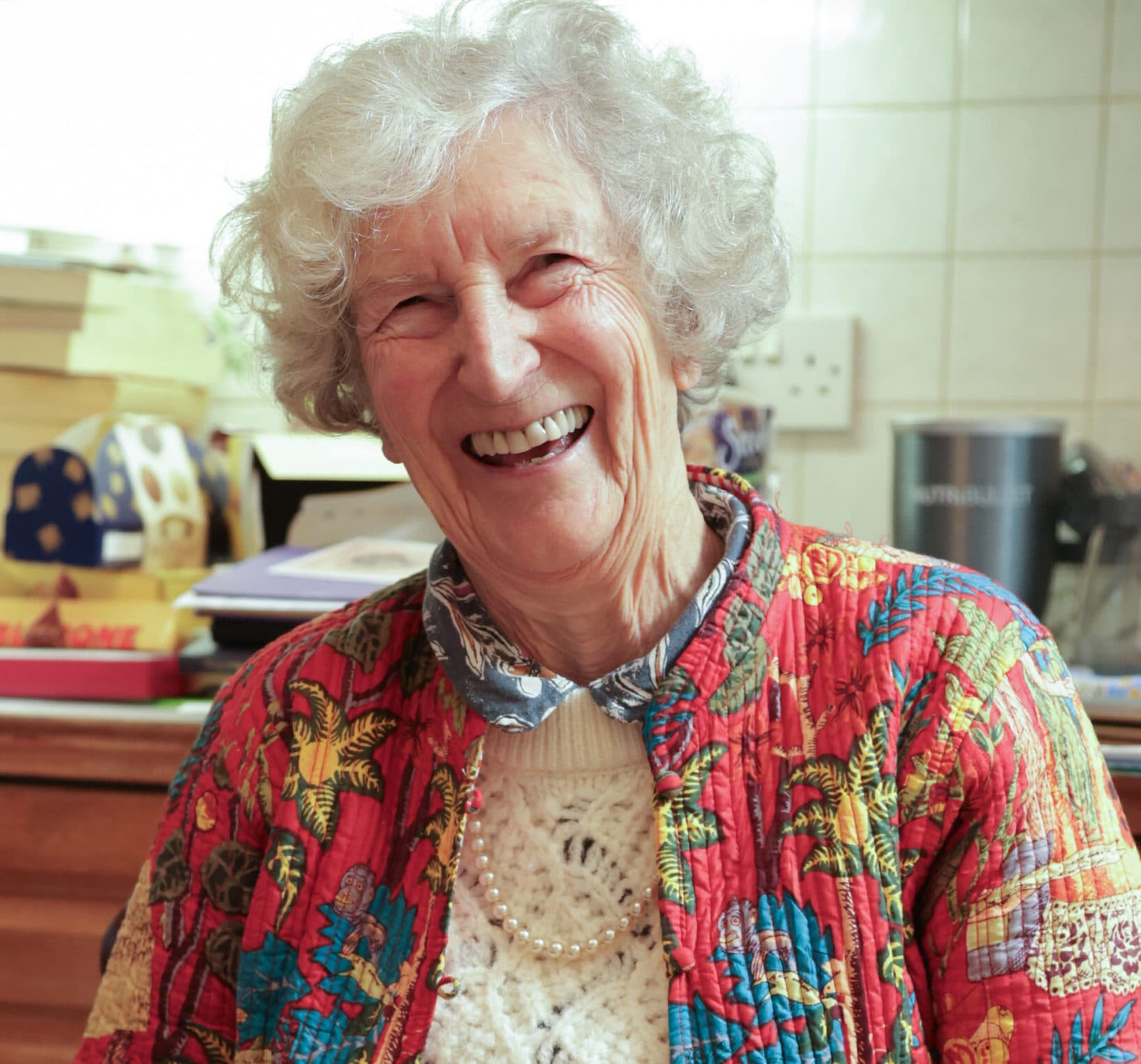 Smiling elderly woman in a colorful floral jacket sits indoors with books and kitchen items in the background. - Home Instead