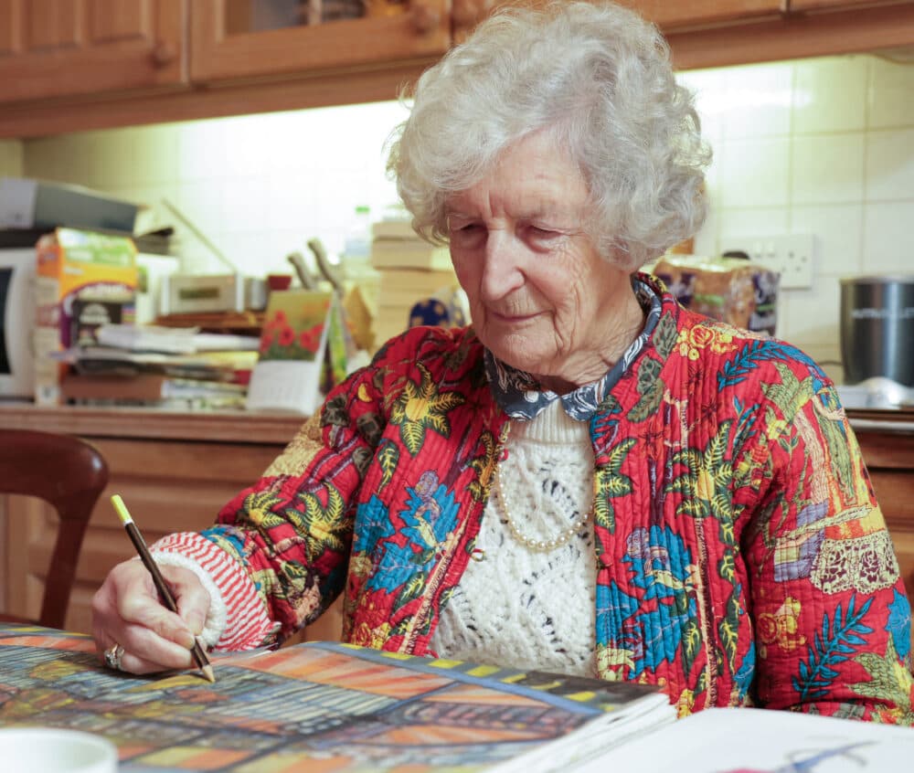 Elderly woman in a colorful jacket drawing at a kitchen table with art supplies. - Home Instead