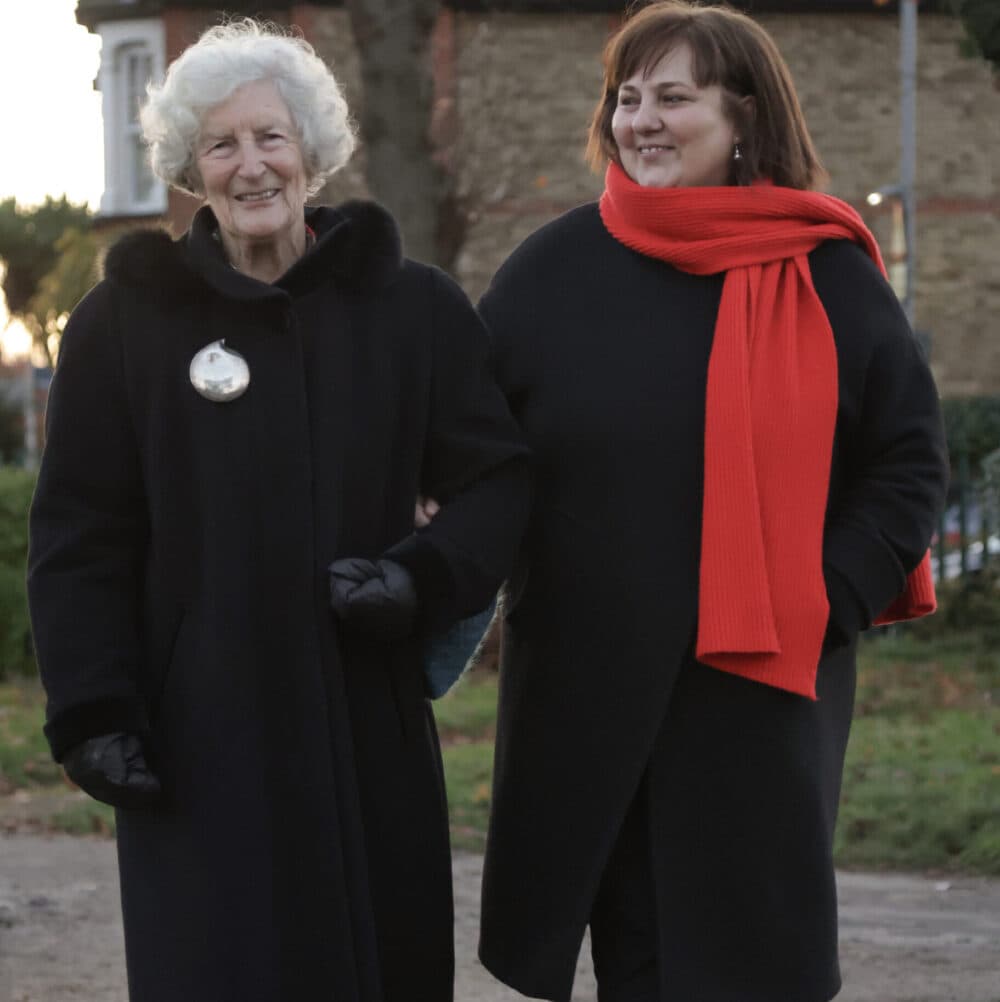 Two women in dark coats walking outside, one wearing a red scarf and both smiling. - Home Instead