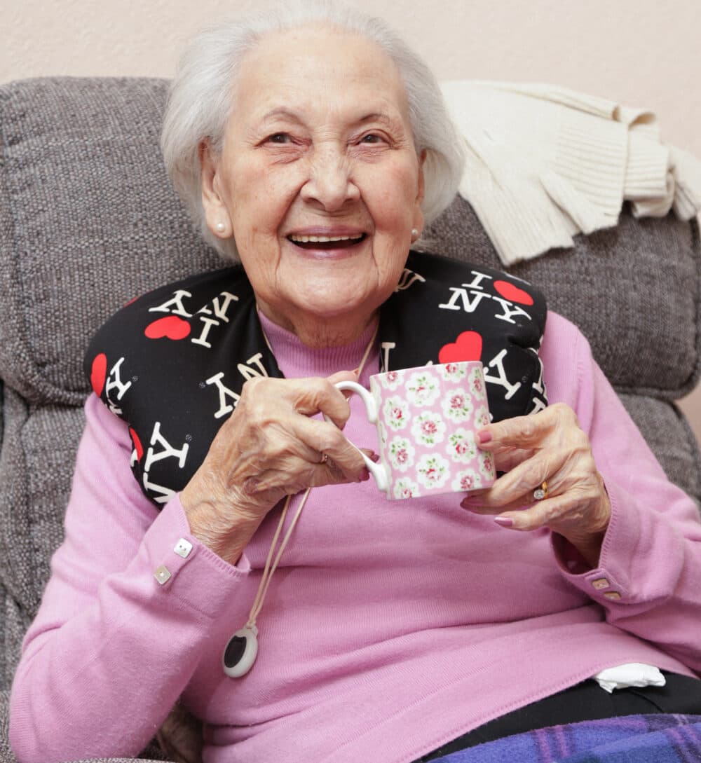 Smiling elderly woman in a pink sweater holding a floral mug, with an "I ♥ NY" neck pillow. - Home Instead