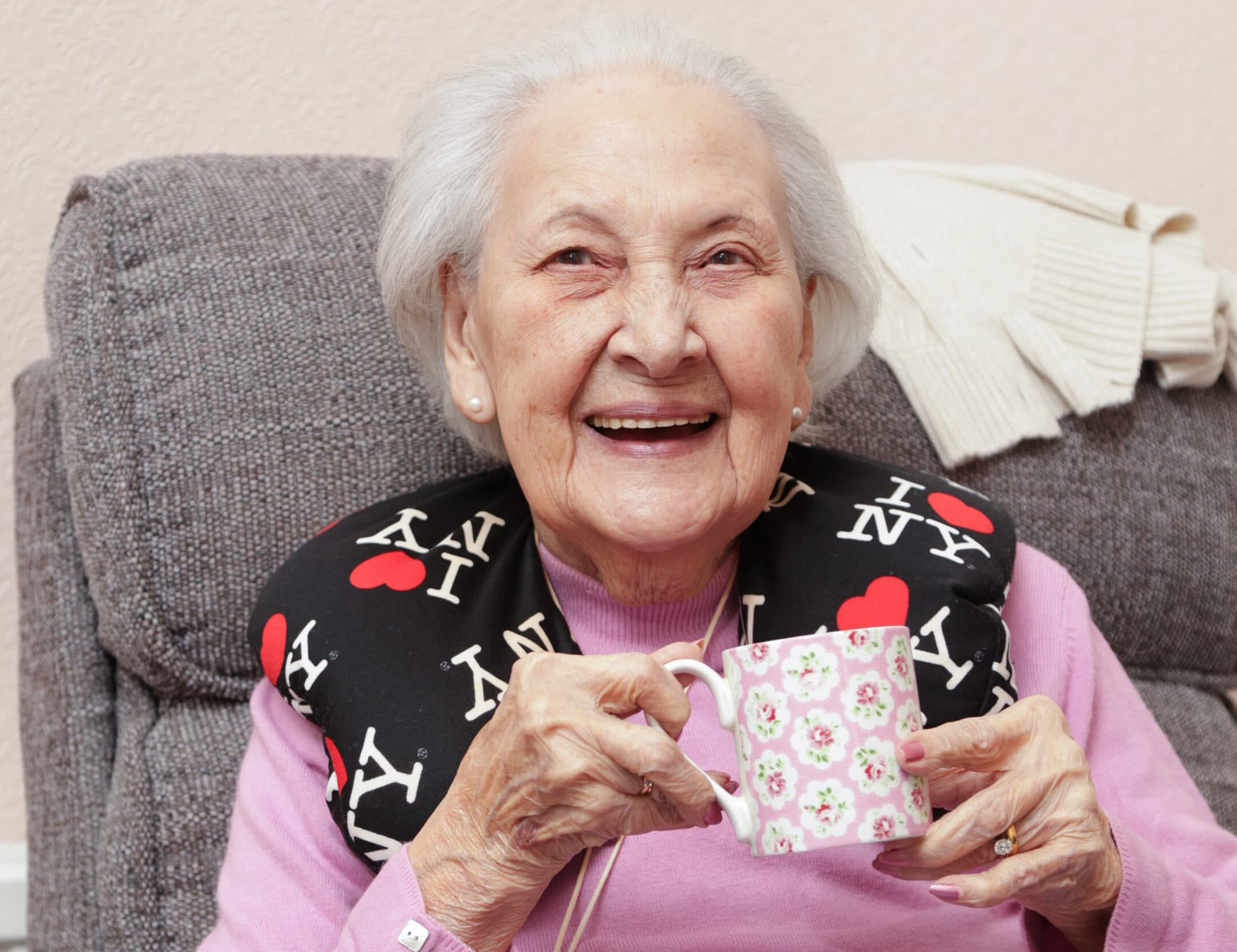 Smiling elderly woman holding a floral mug, wearing a "I ♥ NY" neck pillow while sitting on a gray sofa. - Home Instead