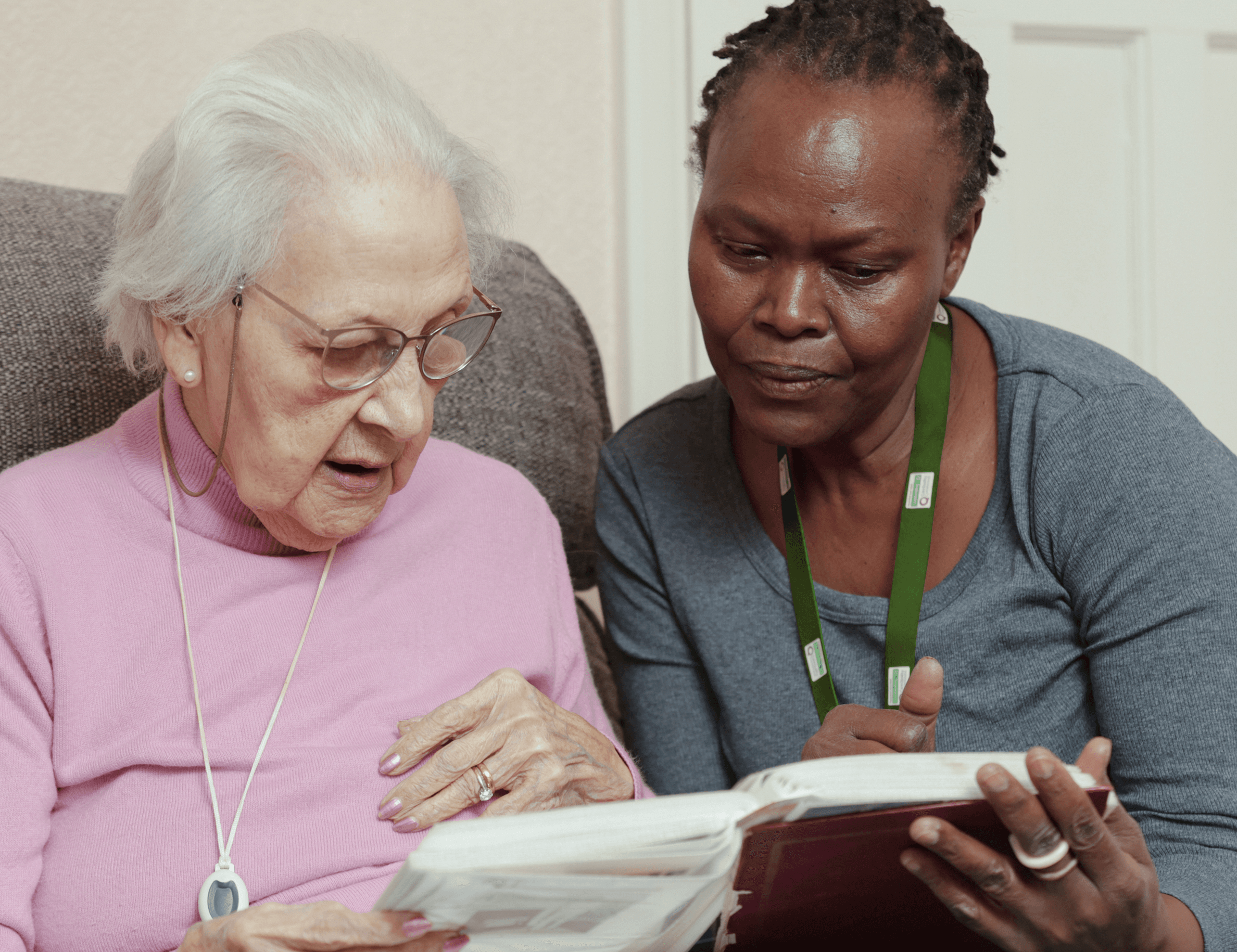 Elderly woman and caregiver looking at a photo album together, sitting closely on a couch. - Home Instead