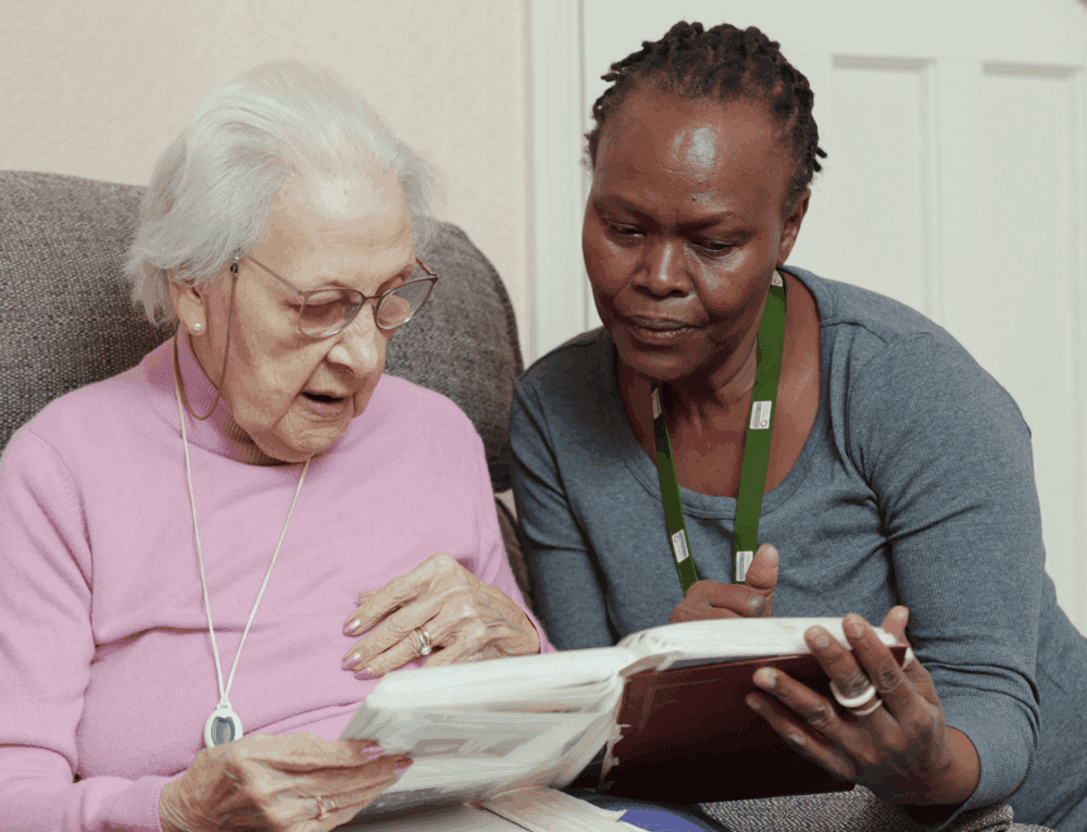 An elderly woman and a caregiver sit together, looking at a photo album and sharing memories. - Home Instead