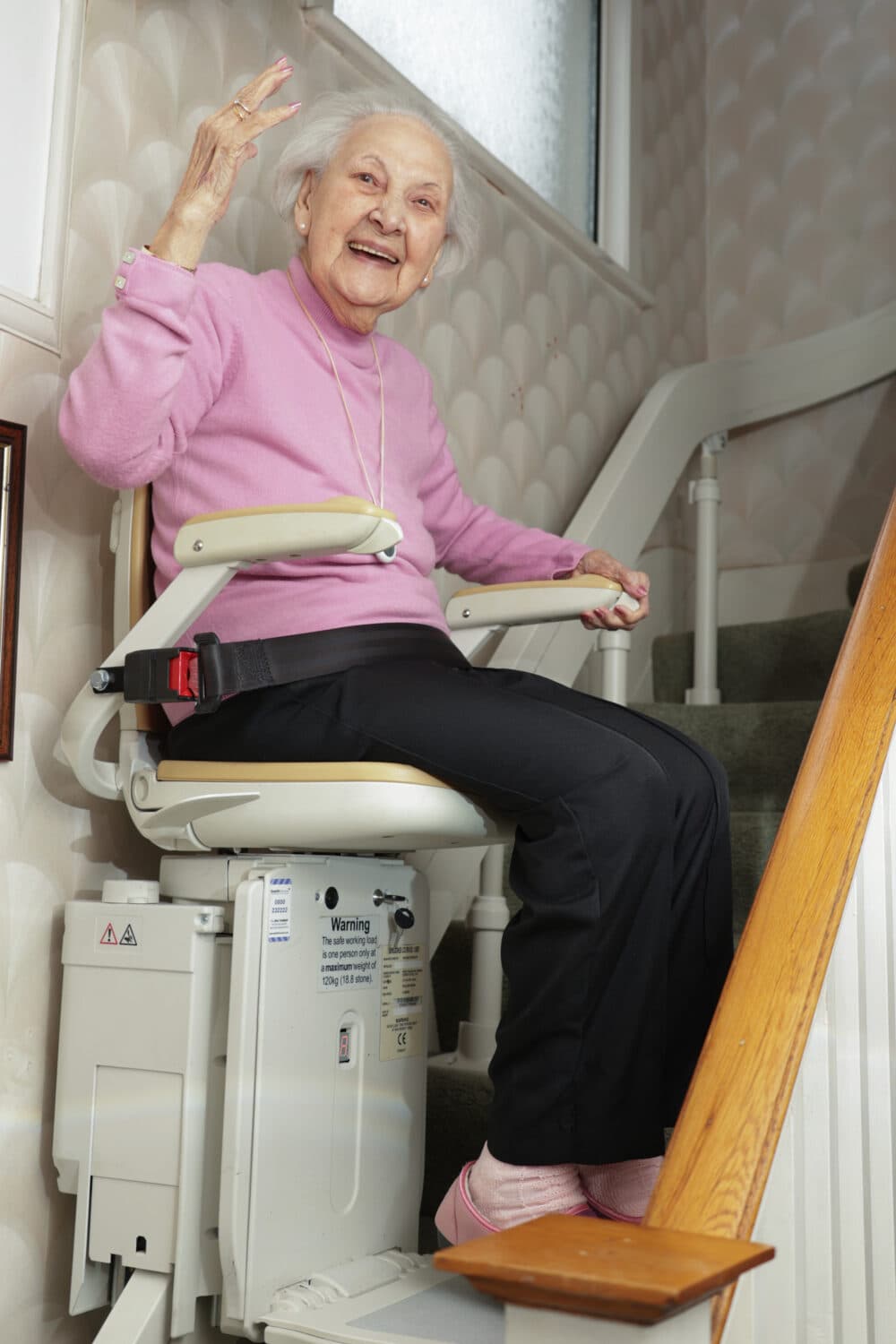 Elderly woman smiling and waving while sitting on a stairlift in a home stairway. - Home Instead