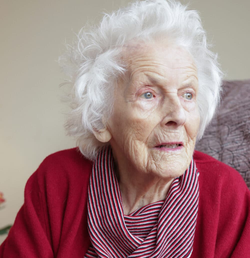 Elderly woman with white hair in a red sweater looking thoughtfully to the side. - Home Instead