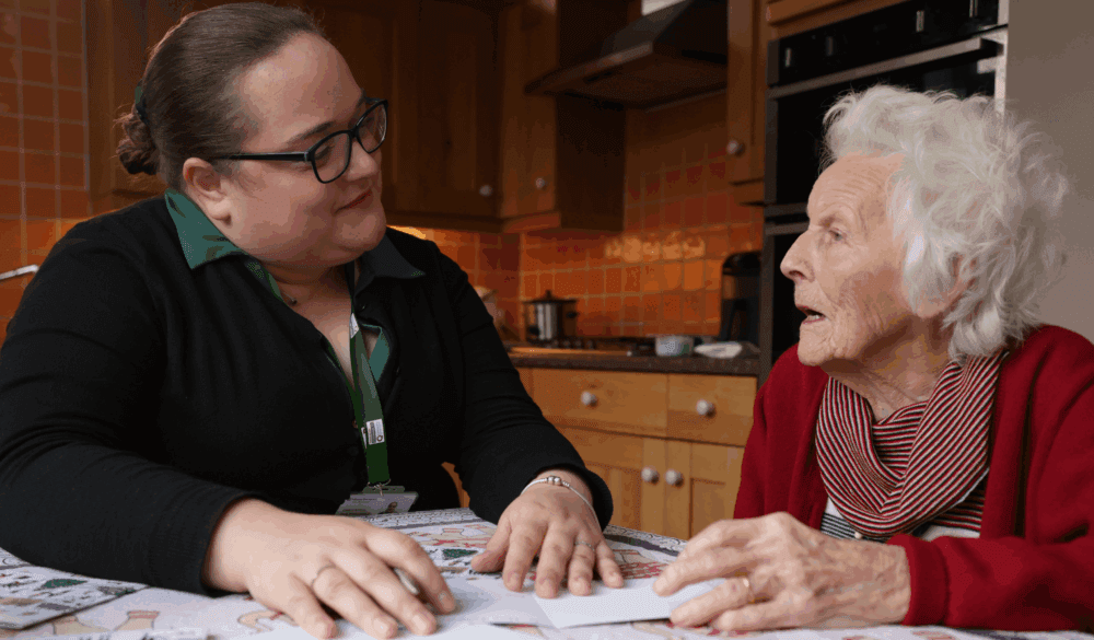 A young woman talks with an elderly woman at a kitchen table, both appearing engaged in conversation. - Home Instead