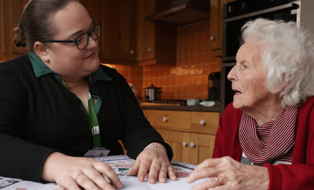 A caregiver sits at a table, talking with an elderly woman in a kitchen. - Home Instead