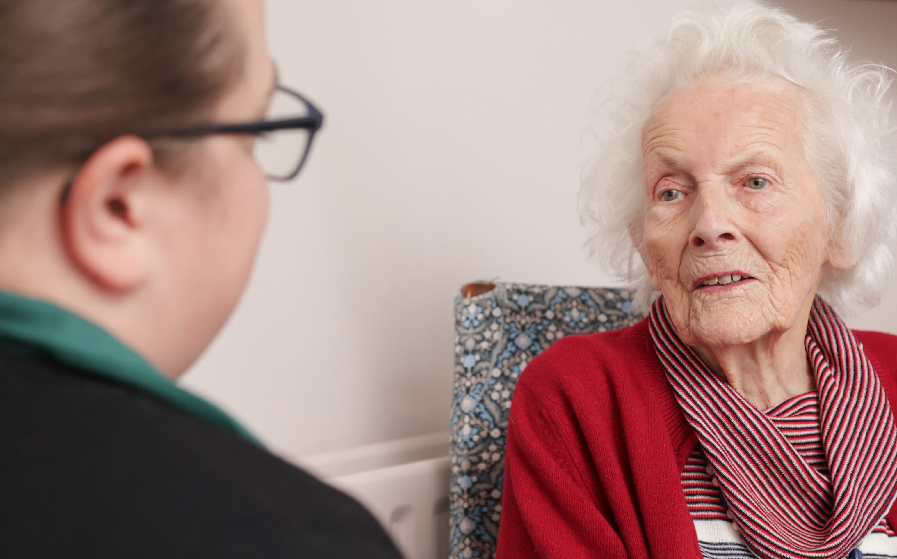 Elderly woman in a red cardigan talking with another person indoors. - Home Instead