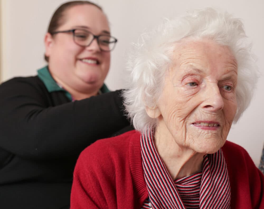 An elderly woman in a red sweater smiles, with a younger woman smiling in the background. - Home Instead
